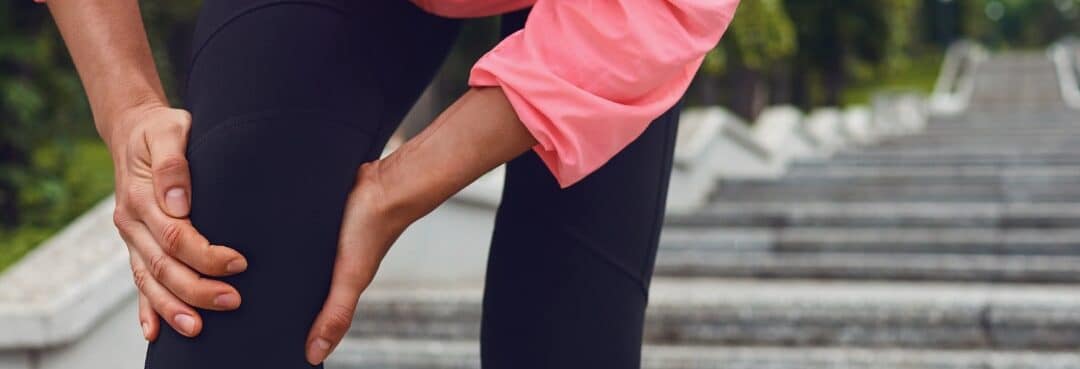 foot ankle leg pain sports injury. you see the bottom half of a torso, both legs bent with both hands around one knee as if they are in pain. the person is wearing pink shoes, black leggings, and a pink long sleeve hoodie.