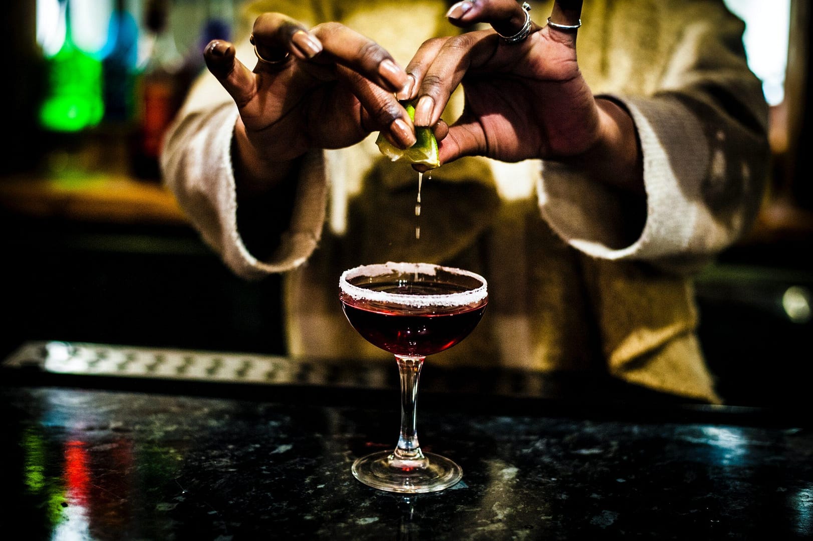 a pair of melanated hands squeezing a lime into a cocktail drink on a dark granite table.