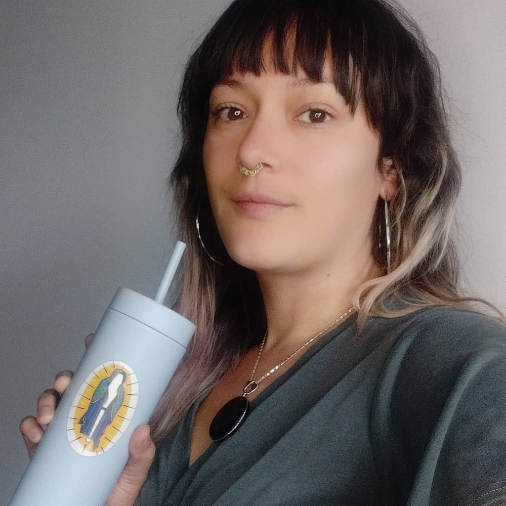 Ashley Stafford holing a blue water cup with a virgin of guadelupe themed juggling club. She has a seagreen top, silver hoop earrings, and an obsidian pendant.