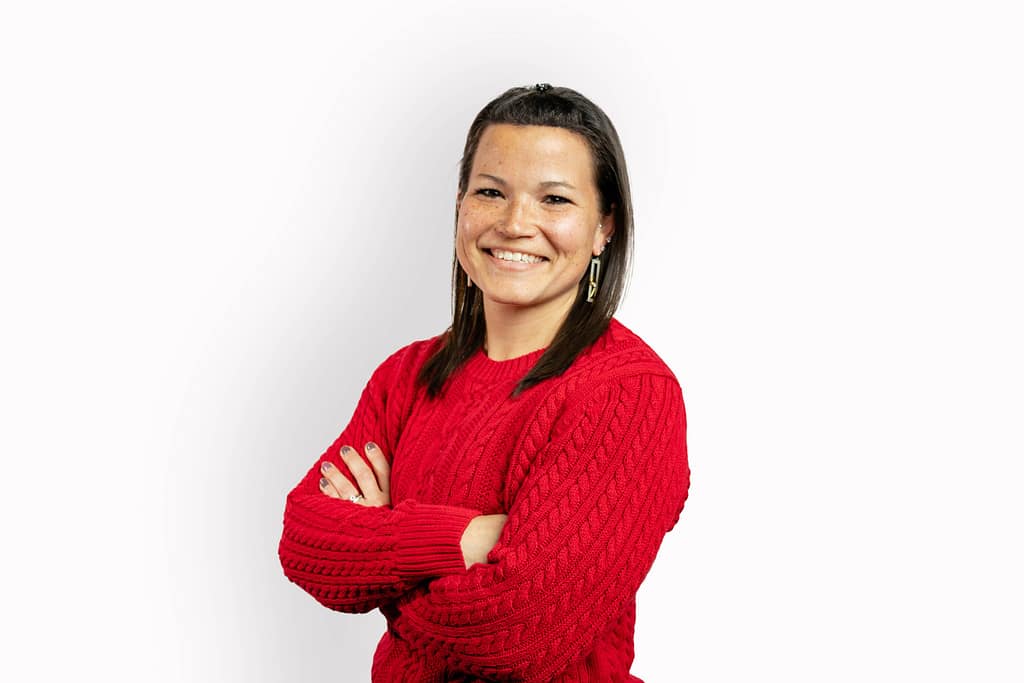 a medium melanated woman smiling looking directly at the camera with her arms crossed. she is wearing a red sweater