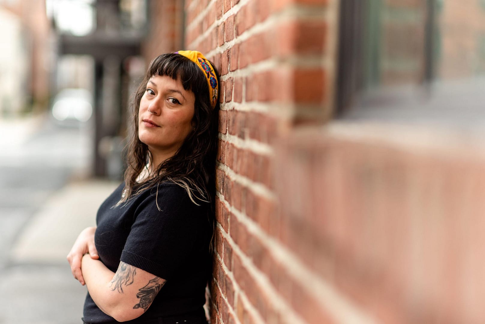 woman facing the left side of the image with her back to a brick wall. She is looking at the viewer.