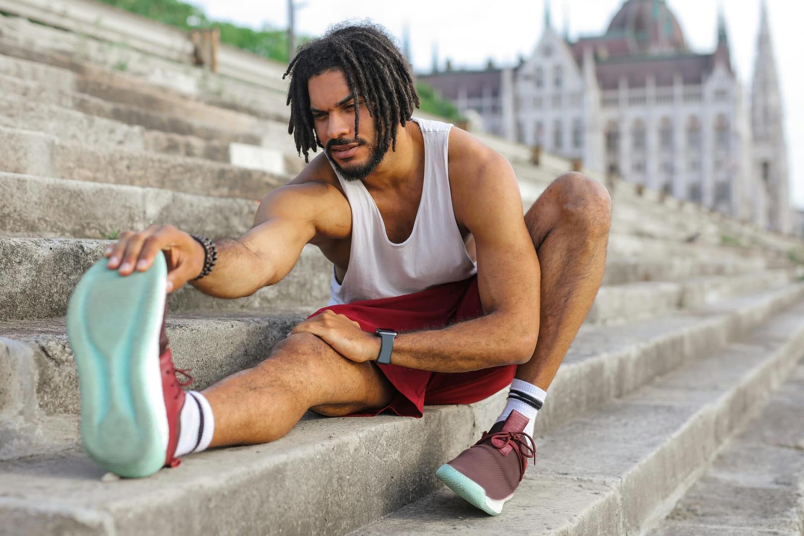 a man is stretching his sciatic nerve on concrete stairs. Wearing red shoes, red shorts, and a white tank. The photo demonstrates sciatica pain relief