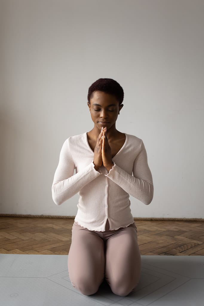 A melanated woman is sitting on her feet with her knees facing the camera. She has her eyes closed and her hands in a praying position. She is wearing a long sleeve, button up, cream colored top with muted coffee colored leggings. She is sitting on a grey yoga mat where you can see the single herringbone wood floor behind her against a white wall.