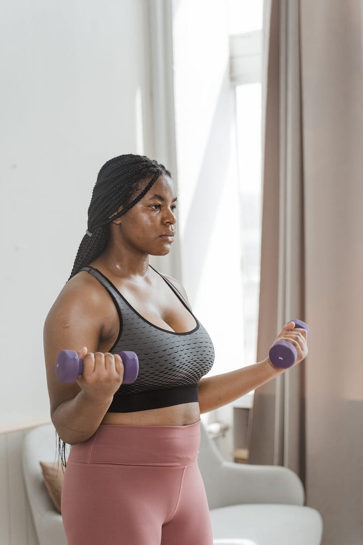 a woman facing the right side of the image holding 3lb weights with her lock ties back in a pony tail. She is wearing a grey sports bra and blush colored leggings.