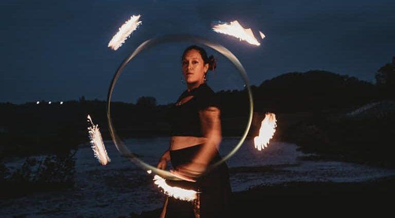 woman dressed in all black facing the left side of the image, looking at the viewer, while holding a flaming hula hoop.