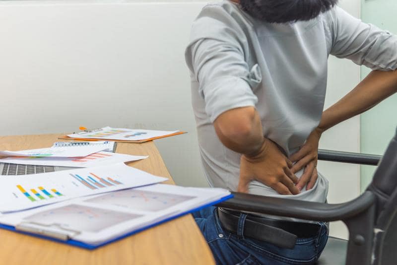 a human with her hands on her low back seated in a chair facing away from the viewer. next to them is a table with paperwork strewn about.